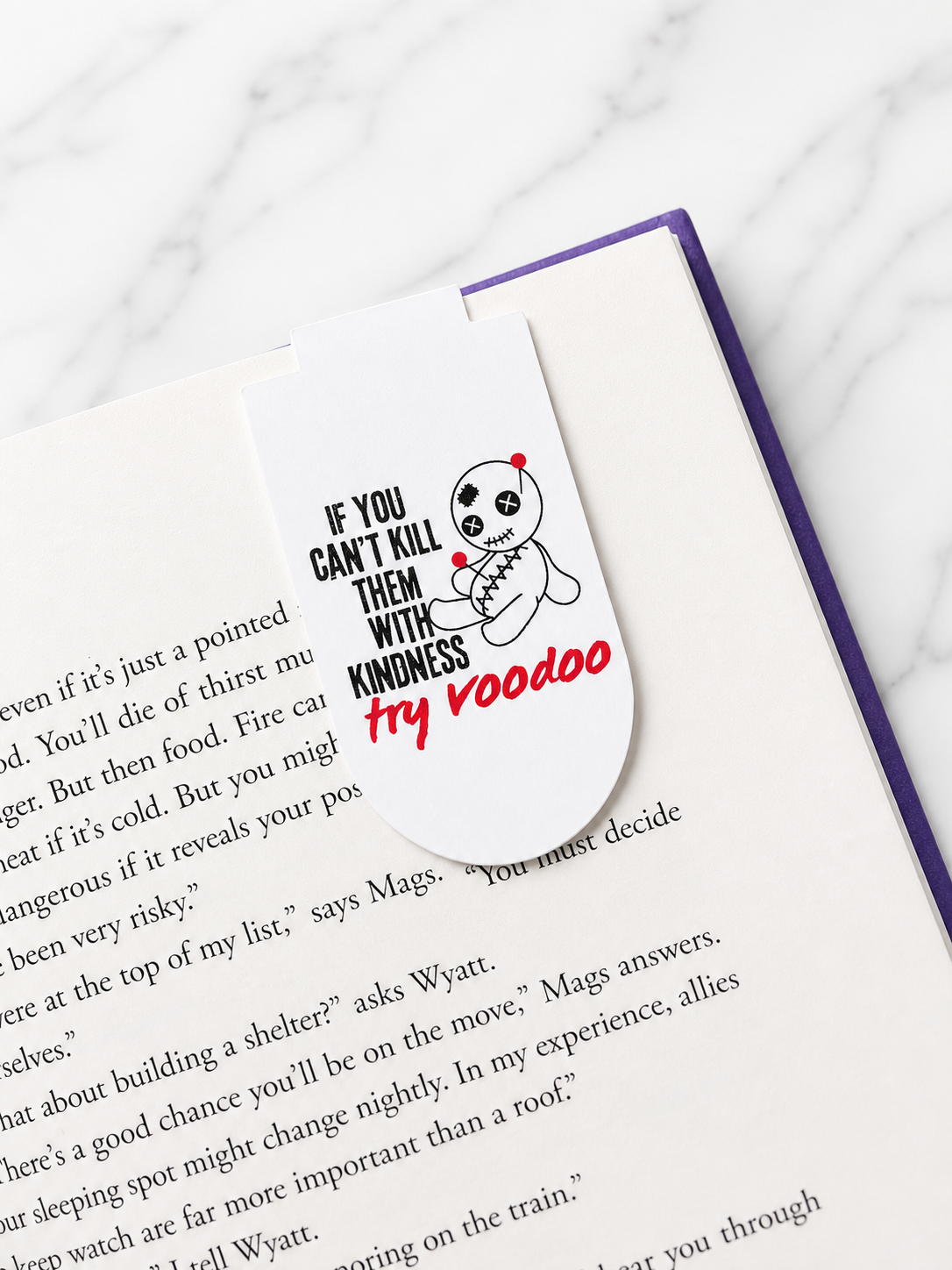 Magnetic bookmark clipped onto the top of an open book page featuring a voodoo doll design and humorous text, demonstrating how it securely marks pages