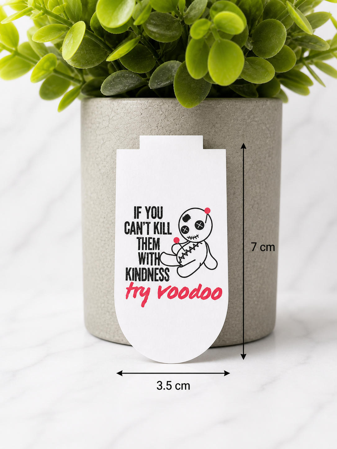 Magnetic bookmark with funny voodoo doll design and the quote “If you can’t kill them with kindness try voodoo”, shown with measurements 7 cm by 3.5 cm on a marble background with plant