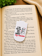 Funny voodoo doll magnetic bookmark resting on an open book in a cosy reading setting with warm lighting, greenery and soft fabric background