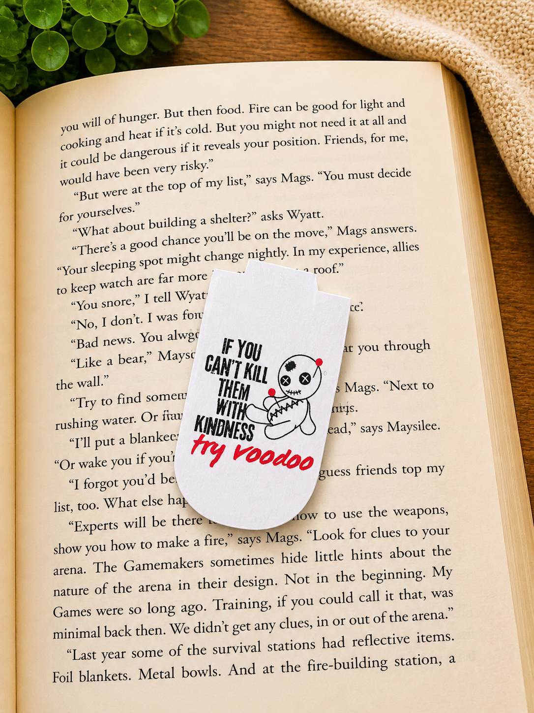 Funny voodoo doll magnetic bookmark resting on an open book in a cosy reading setting with warm lighting, greenery and soft fabric background