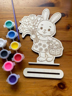 close up of a wooden cutout of a rabbit holding flowers with paint containers and a paintbrush on a wooden surface. Below it is a stand you can put your finish design on.