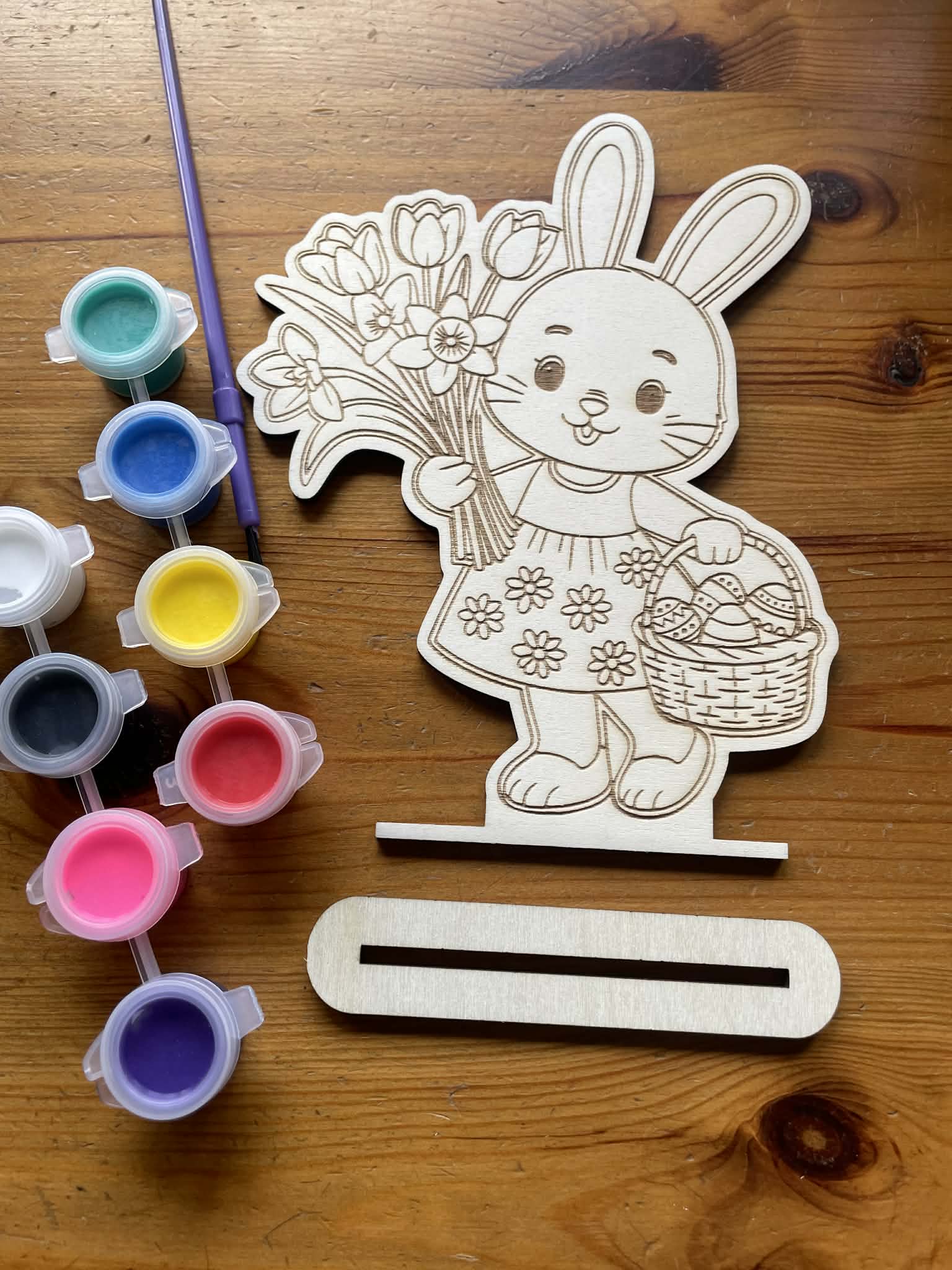 close up of a wooden cutout of a rabbit holding flowers with paint containers and a paintbrush on a wooden surface. Below it is a stand you can put your finish design on.