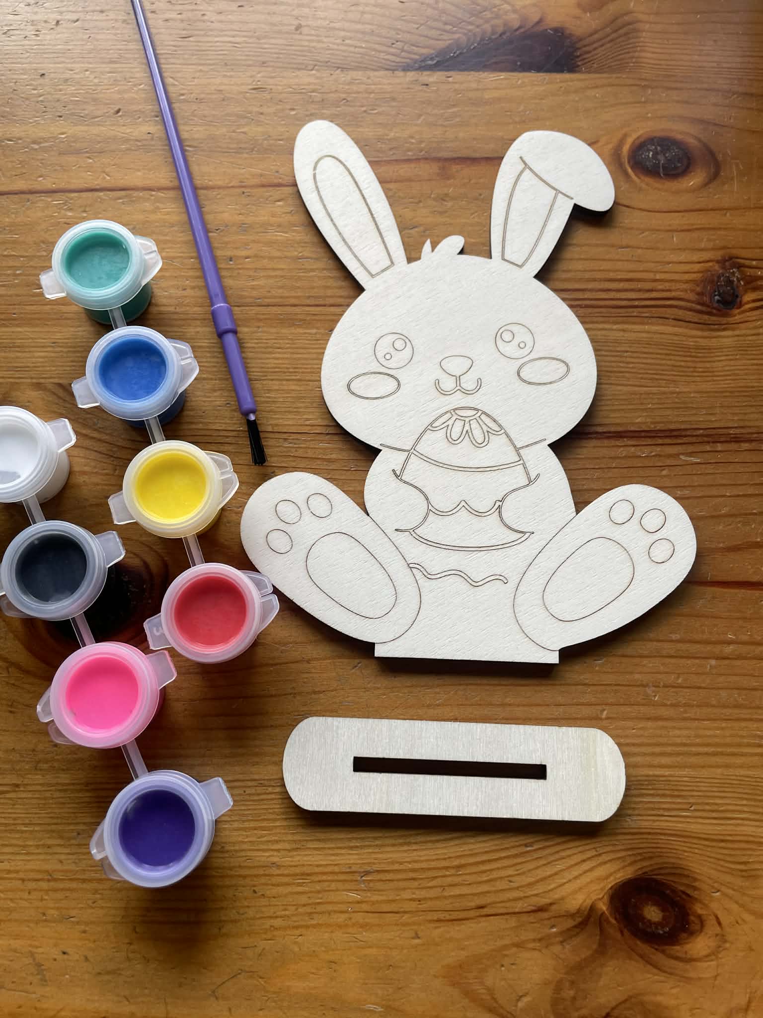 close up of a wooden cutout of a bunny holding an easter egg with paint containers and a paintbrush on a wooden surface. Below it is a stand you can put your finish design on.