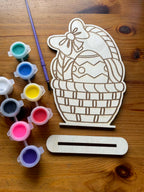 close up of a wooden cutout of a basket of easter eggs with paint containers and a paintbrush on a wooden surface. Below it is a stand you can put your finish design on.