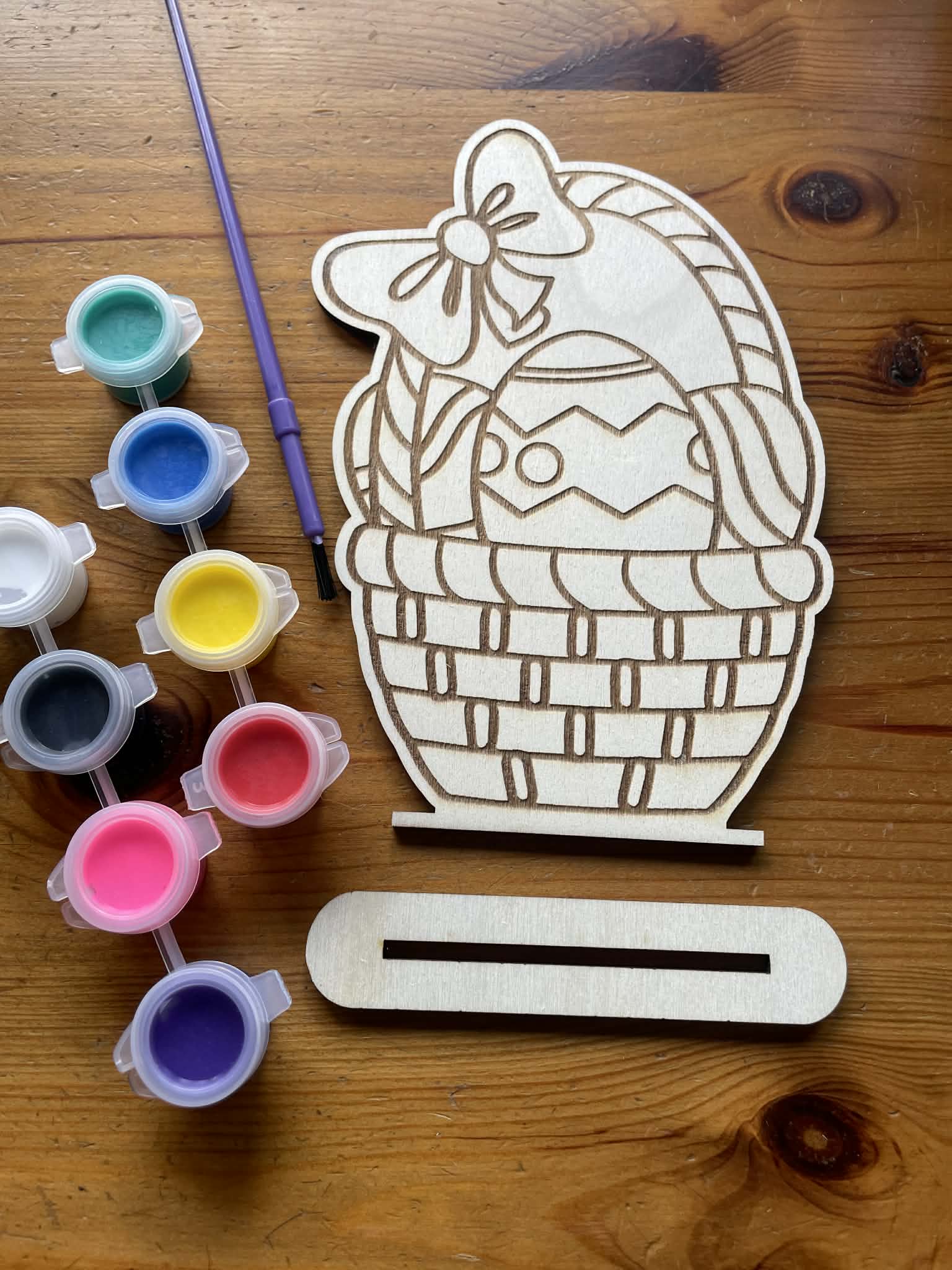 close up of a wooden cutout of a basket of easter eggs with paint containers and a paintbrush on a wooden surface. Below it is a stand you can put your finish design on.