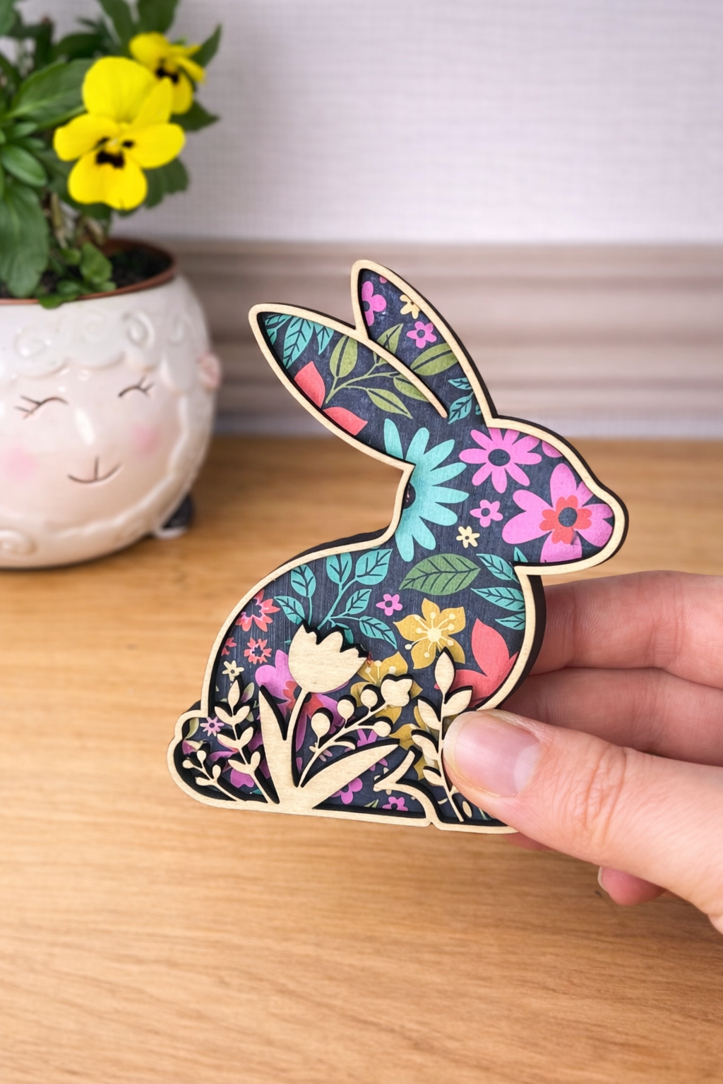 A hand holding a decorative rabbit figurine with floral patterns on a dark background and a cut out of flowers following the bottom silhouette of the rabbit. 