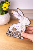 A hand holding a decorative rabbit figurine with floral patterns on a white background and a cut out of flowers following the bottom silhouette of the rabbit. The silhouette is stained a dark brown for a rustic look.
