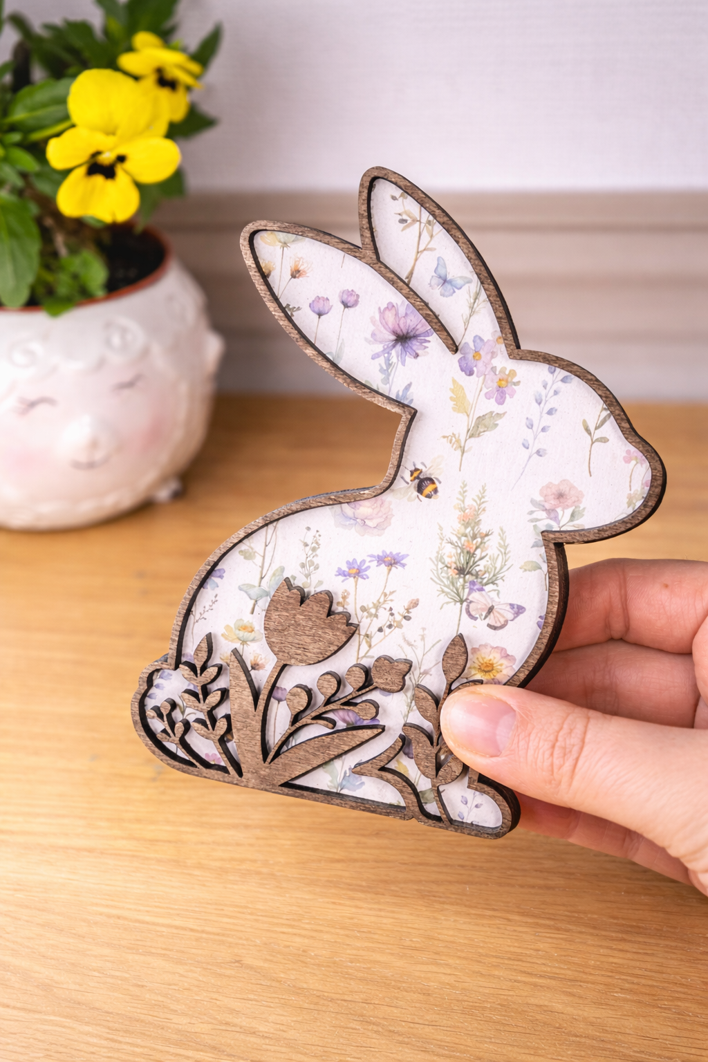 A hand holding a decorative rabbit figurine with floral patterns on a white background and a cut out of flowers following the bottom silhouette of the rabbit. The silhouette is stained a dark brown for a rustic look.