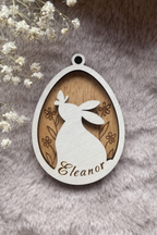 Wooden tag with a bunny and butterfly inside an egg with floral background stained teak/ light brown. On the bottom is space for a personalised name.
